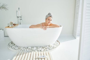 Genç, sarışın, köpüklü, kafasında havlu olan çekici bir kadın rahatlatıcı bir banyo yapar, eğlenir ve gülümser. Ev spası, beyaz otel banyosu. İç mekan.