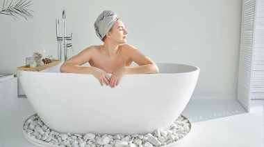 Genç, sarışın, köpüklü, kafasında havlu olan çekici bir kadın rahatlatıcı bir banyo yapar, eğlenir ve gülümser. Ev spası, beyaz otel banyosu. İç mekan.