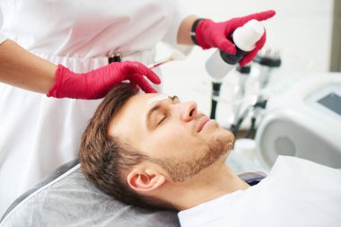 Güzellik salonundaki kozmetoloji prosedürlerinde çalışan yakışıklı beyaz bir genç adam. Erkek dermatolog cilt bakımı, yüz maskesi.