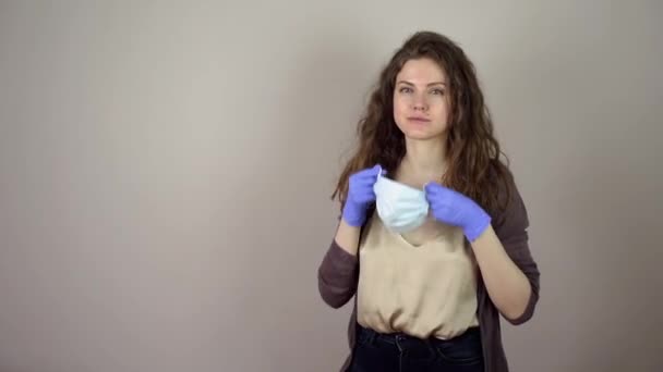 Brunette jonge vrouw zetten beschermende masker op gezicht in coronavirus pandemie op grijze achtergrond. — Stockvideo