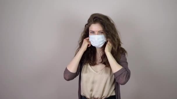 ผมสีบรูเนทหญิงสาวถอดหน้ากากป้องกันบนใบหน้าในการระบาดของไวรัสโคโรนาบนพื้นหลังสีเทา หายใจและยิ้ม — วีดีโอสต็อก