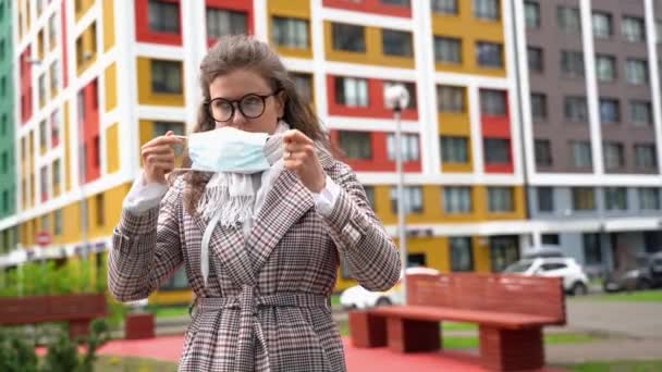 Jeune belle femme caucasienne en lunettes met un masque blanc sur son visage pour se protéger pendant une pandémie — Video