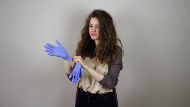 Brunette young woman putting on blue gloves in coronavirus pandemic on grey background — Stock Video