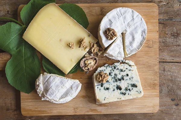 Various french cheeses, nuts, fig leaf on the wooden cutting board — Stock Photo, Image