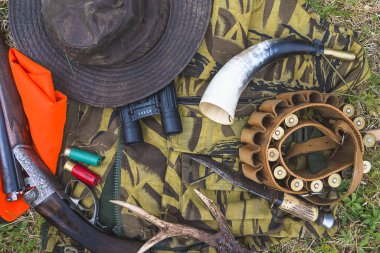 Avlanma teçhizatı üst görünümde. Av silahı, kemer fişekleri, şapka, dürbün, av boynuzu, geyik boynuzu, bıçak.