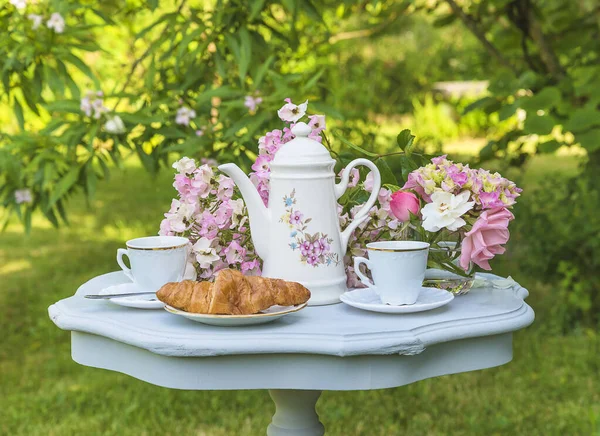 Coffee break in blooming summer garden. Vintage coffee pot, two cups of coffee, croissant and bunch of rose flowers on vintage table in the garden
