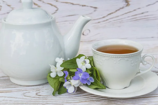 Tea time concept in retro style. White cup of tea, tea pot and bouquet of violets