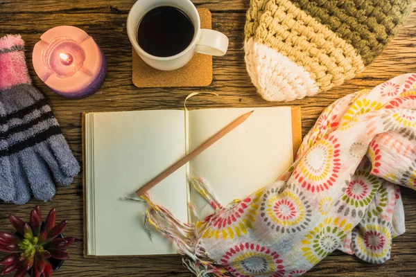 Fincan kahve, defter, mumlar, şapka, eşarp ve eldiven ahşap bir masa üzerinde.