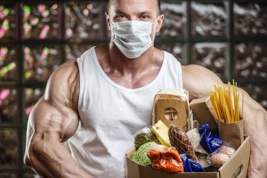 Baltalı vücut geliştirici sporcu, spor salonunda gaz maskeli yemek. Peynirli yumurta ve mısır gevreğiyle kaslı adam Coronavirüs salgını covid-19 pandemik kaos medikal maske kıyamet sonrası vücut geliştirme.