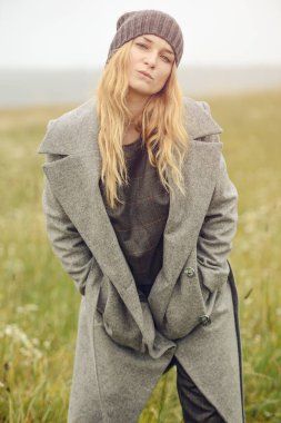 Güzel giyimli, güzel, beyaz, sarı saçlı, şık elbiseli, genç bir kız. Paltolu, açık hava, çiftlik evinin yanında, kırsal alanda, bahar veya sonbahar havası depresif.