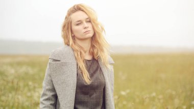 Güzel giyimli, güzel, beyaz, sarı saçlı, şık elbiseli, genç bir kız. Paltolu, açık hava, çiftlik evinin yanında, kırsal alanda, bahar veya sonbahar havası depresif.