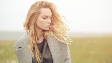 Güzel giyimli, güzel, beyaz, sarı saçlı, şık elbiseli, genç bir kız. Paltolu, açık hava, çiftlik evinin yanında, kırsal alanda, bahar veya sonbahar havası depresif.