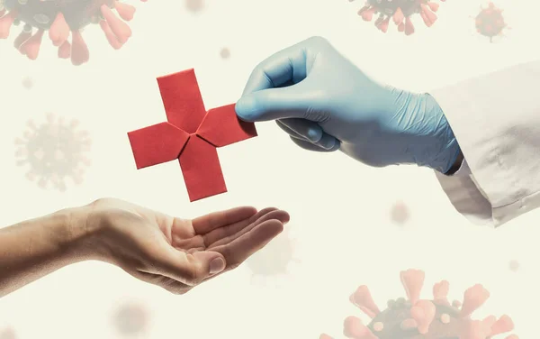 Doctor\'s hand gives a red paper cross  to a woman. Image on background with coronavirus covid-19. Concept of medical treatment during the pandemic covid-19