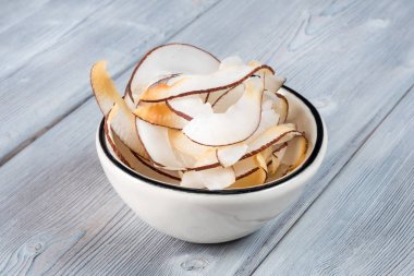 Beyaz ahşap arka planda, beyaz kasede kurutulmuş hindistan cevizi parçaları.