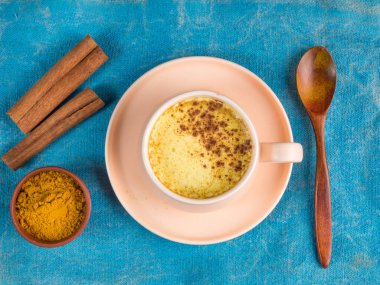 Üzerinde zerdeçal, tarçın ve ahşap kaşık olan bir fincan altın sütün mavi arka planda görüntüsü.