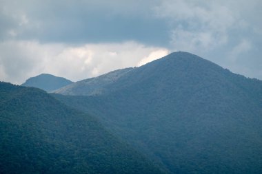 Bulutlu bir gökyüzünün altında, bulutlardaki bir delikten parlayan güneş ve dağda yanan bir nokta yapan bir dağ..
