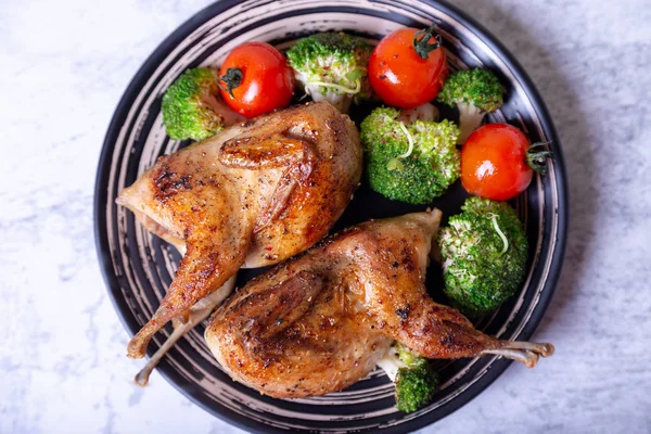 stock image Quail fried whole with broccoli and cherry tomatoes. Black plate, marble background. Selective focus, close up.