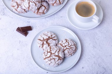 Çatlaklı çikolatalı kurabiyeler, üzerine şeker serpiştirilmiş. Ev yapımı pişirme. Arka plan: bir fincan kahve, çikolata ve bir tabak kurabiye. Seçici odak, yakın plan.