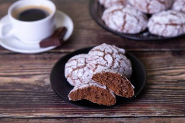 Çatlaklı çikolatalı kurabiyeler, üzerine şeker serpiştirilmiş. Ev yapımı pişirme, ahşap arka plan. Arka plan: bir fincan kahve, çikolata ve bir tabak kurabiye. Seçici odak, yakın plan.