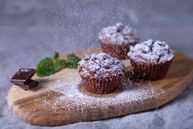 Tahtaya çikolatalı kek serpilmiş, krema şekerli. Ev yapımı pişirme. Arka planda bir fincan kahve ve bir tabak kek var. Ahşap arka plan. Seçici odak, kapat.