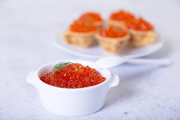 Red caviar (salmon caviar) in a white cup. In the background is a plate with tartlets and caviar. Selective focus, close up.