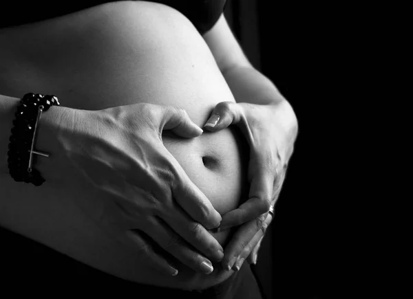 Mujer embarazada sosteniendo sus manos en forma de corazón sobre su protuberancia aislada sobre fondo negro. Embarazada vientre con símbolo del corazón. Concepto de maternidad — Foto de Stock