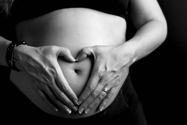 Zwangere vrouw hand in haar hand in de vorm van een hart op haar Baby Bump geïsoleerd op zwarte achtergrond. Zwangere buik met hartsymbool. Zwangerschaps Concept — Stockfoto