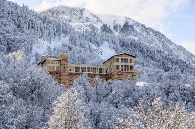 Beautiful Solis Sochi Hotel surrounded with snowy forest trees on a sunny mountain slope background scenic landscape clipart