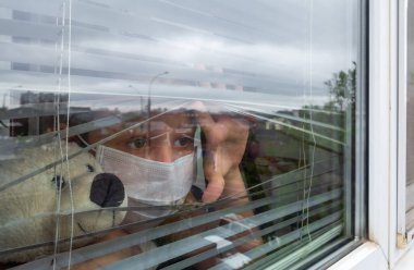 Evde kalıp koronavirüs salgını konseptini karantinaya alın. Koruyucu tıbbi maskeler takan, elinde oyuncak ayıyla pencereden dışarı bakan üzgün bir kız. Salgın önleme kavramı