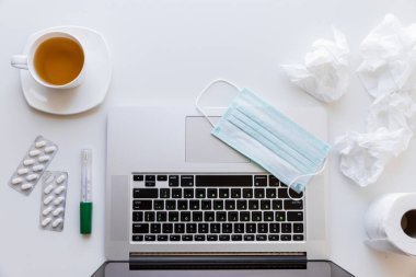 Karantinada bir fincan çay ve çay tabağı, klavyede tıbbi maske, bir rulo tuvalet kağıdı, peçete, tablet ve termometre kullanılmış bir laptop. Bunların hepsi beyaz bir masanın üzerinde.