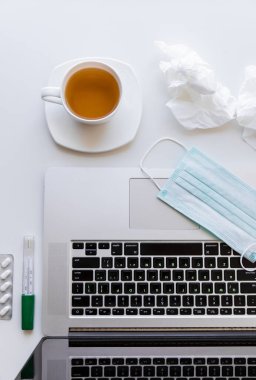 Karantinada bir fincan çay ve çay tabağı, klavyede tıbbi maske kullanılmış peçeteler, tabletler ve termometre olan bir laptop. Bunların hepsi beyaz bir masanın üzerinde.