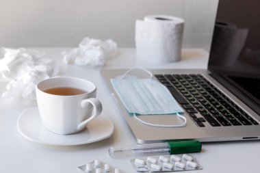 Karantinaya alınmış bir fincan çay ve çay tabağı, klavyede tıbbi maske, bir rulo tuvalet kağıdı ve kullanılmış peçeteler, tabletler ve termometre. Bunların hepsi beyaz bir masanın üzerinde.