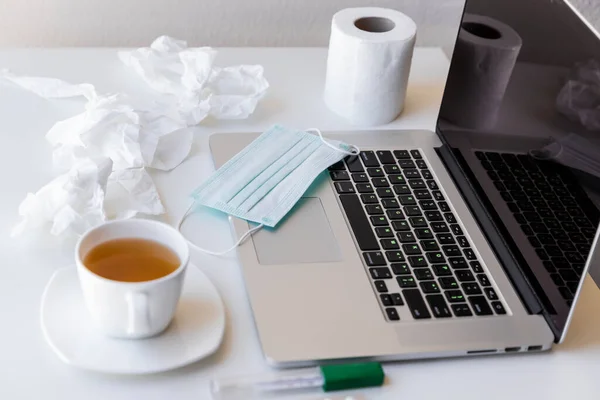 Karantinada bir fincan çay ve çay tabağı olan bir laptop klavyede tıbbi maske bir rulo tuvalet kağıdı ve kullanılmış peçeteler, termometre. Her şey beyaz masada.