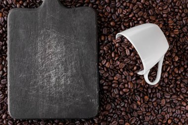 Beyaz bir kupa ve her yerde kavrulmuş kahve çekirdeklerinin yanında gri tahta bir tahta var. Kupanın içinde ve etrafında bir sürü şey var. Kafeye, restorana, kafeteryaya ya da tabela olarak gitmek için harika.