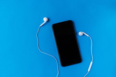 Mavi arka planda beyaz kulaklıklı, koyu ekran kapaklı modern bir cep telefonu. Güzel duvar kağıdı. Popüler bir resim. Üst Manzara. Aktif gençler için yaşam tarzı