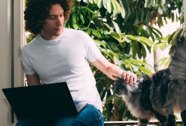 Beyaz tişörtlü, kıvırcık saçlı genç bir adam elinde dizüstü bilgisayarla pencerede oturuyor ve Coronavirus karantina döneminde evden çalışıyor. Kediyi okşuyor. Pencerenin dışında yeşil palmiye ağaçları