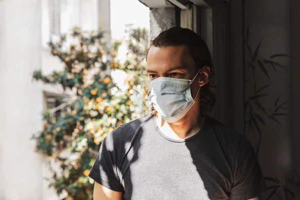 Young man with long hair in a medical mask sits near a window against a green tree with orange fruits. Quarantine during the coronavirus epidemic, covid-19. Waiting for the end of pandemic.