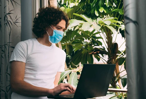 Young curly-haired man in a medical mask and a white T-shirt orders food delivery online from a laptop. He stands near a window from which green trees are visible. Stay home. Gray curtains. Copy space