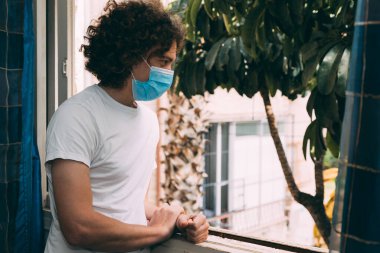 Tıbbi maskeli, beyaz tişörtlü, kıvırcık saçlı genç bir adam karantina altındaki evin penceresinden koronavirüsten dışarı bakıyor. Evde kal konsepti. Covid-19. Tek başıma, iyi haberleri bekliyorum.
