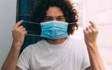 Beyaz tişörtlü bir adam önünde tıbbi bir maske tutuyor, önündeki kayışlardan tutuyor. Koronavirüs salgını sırasında ev karantinasında. Evde kal konsepti. Yeni gerçeklik.