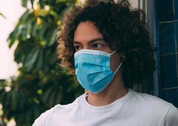 Mavi tıbbi maske ve beyaz tişört giyen kıvırcık uzun saçlı genç bir adamın portresi. Pencerenin yanındaki yeşil arka plan ve mavi perde. Ev karantinası, koronavirüs salgını. Yeni gerçeklik. Evde kal.