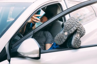 Tıbbi maskeli bir adam, bacakları yukarıda arabada oturuyor. Koruyucu maskeli adam arabadaki cep telefonundan selfie çekiyor. Koronavirüs salgınından korunma, kovid-19. Yeni gerçeklik