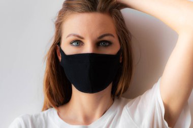 Siyah tıbbi maskeli, mavi gözlü ve beyaz arka planda beyaz tişörtlü genç bir kadının yakın plan portresi. Karantina, koronavirüs salgını, covid-19. Evde kal konsepti.