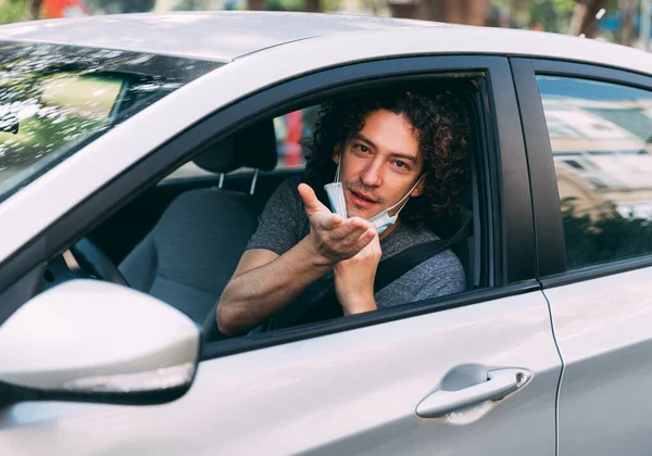 A handsome man with a medical mask driving a car, blows a kiss. A man with a protective mask is sitting behind the wheel of a car. Protection from coronavirus epidemic pandemic, covid-19. New reality