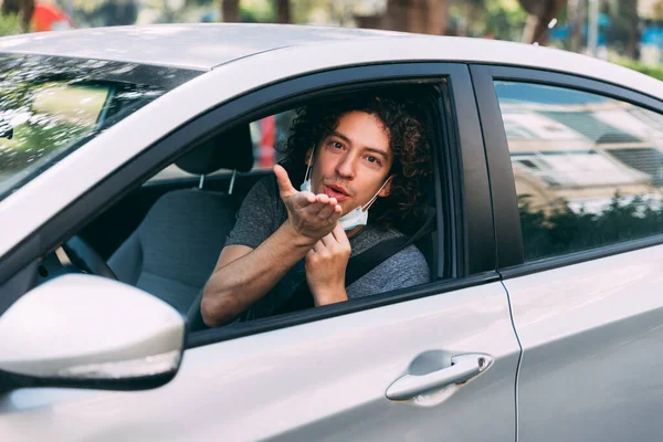 A handsome man with a medical mask driving a car, blows a kiss. A man with a protective mask is sitting behind the wheel of a car. Protection from coronavirus epidemic pandemic, covid-19. New reality