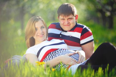 ilkbaharda yeşil çimenlerin üzerinde oturan genç hamile Çift 