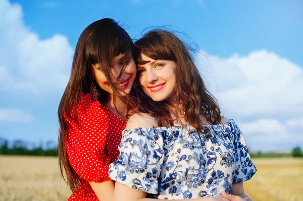 Two cheerful women in a wheat field at sunset in a blue and red long air dress. — Stock Photo, Image