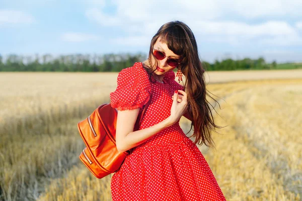 Mulher em um campo de trigo em um vestido longo vermelho, com uma mochila . — Fotografia de Stock