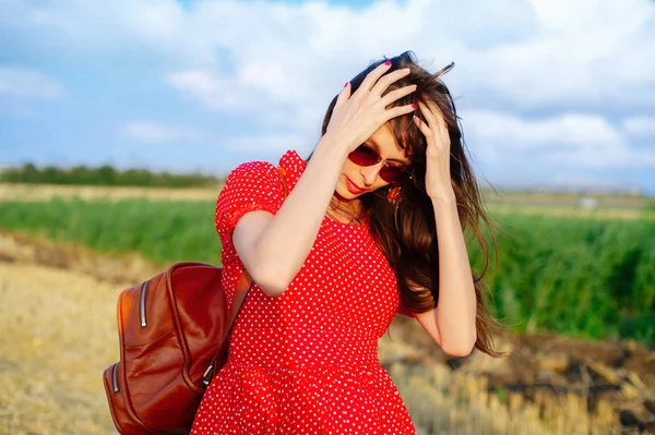 Una mujer sostiene su cabeza, con un vestido largo rojo y una mochila, en un campo, tiempo soleado brillante . — Foto de Stock
