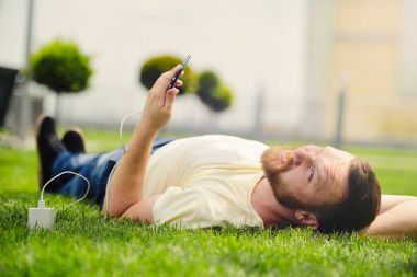 Doğa ve teknoloji. Bir sakallı bir adam yatıyor bir yeşil çim onun elinde bir akıllı telefon ile güç şarj banka kameraya benziyor.